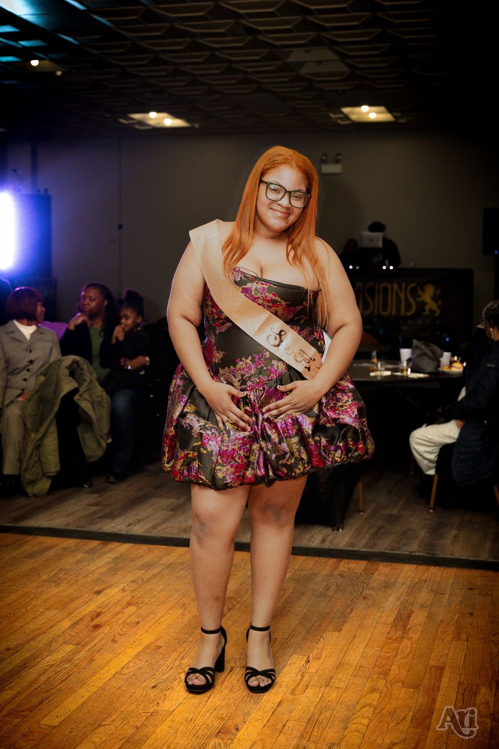Woman in floral dress and sash poses on wooden runway at indoor pageant event.
