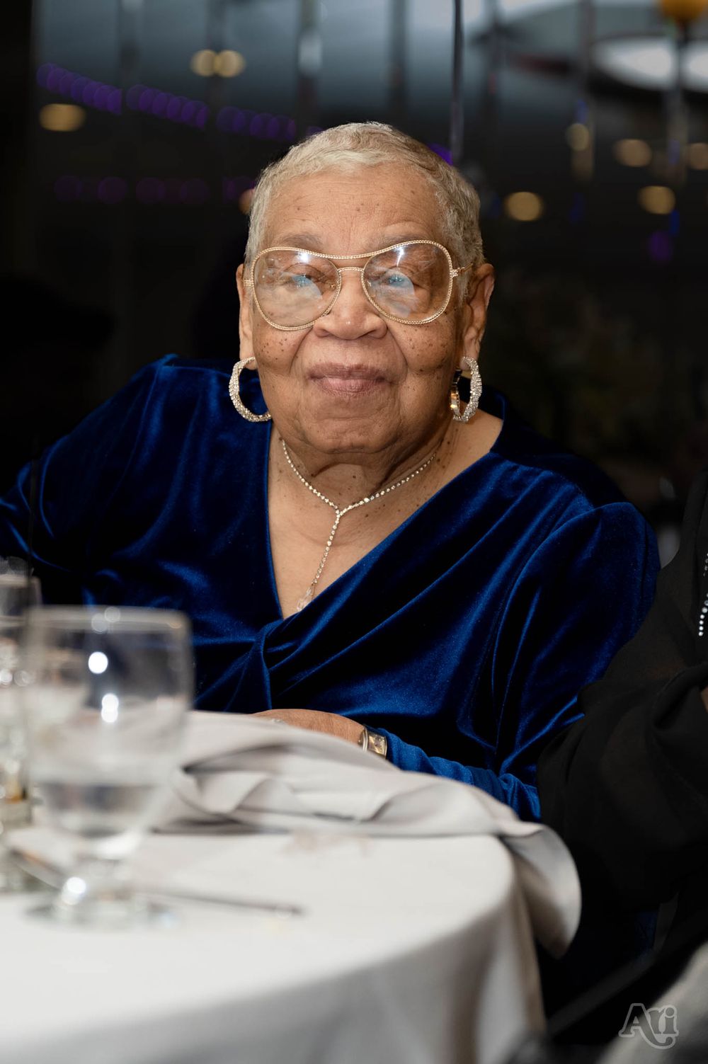 Person in elegant blue velvet dress seated at formal dining table smiles warmly.