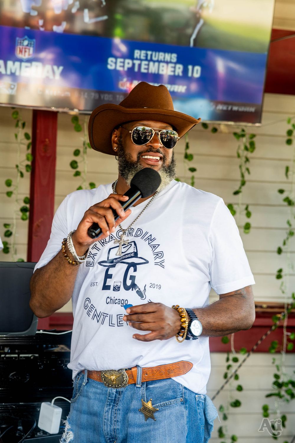 A speaker in a white t-shirt, brown cowboy hat and sunglasses holds a microphone outdoors in front of an NFL GameDay banner.