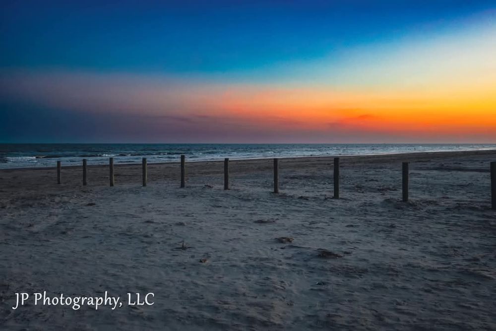 Dramatic sunset over a sandy beach with silhouettes of wooden posts along the shoreline.
