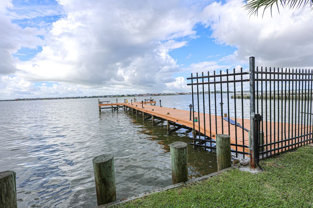 Long wooden pier with metal fence extending into calm waters under cloudy sky.