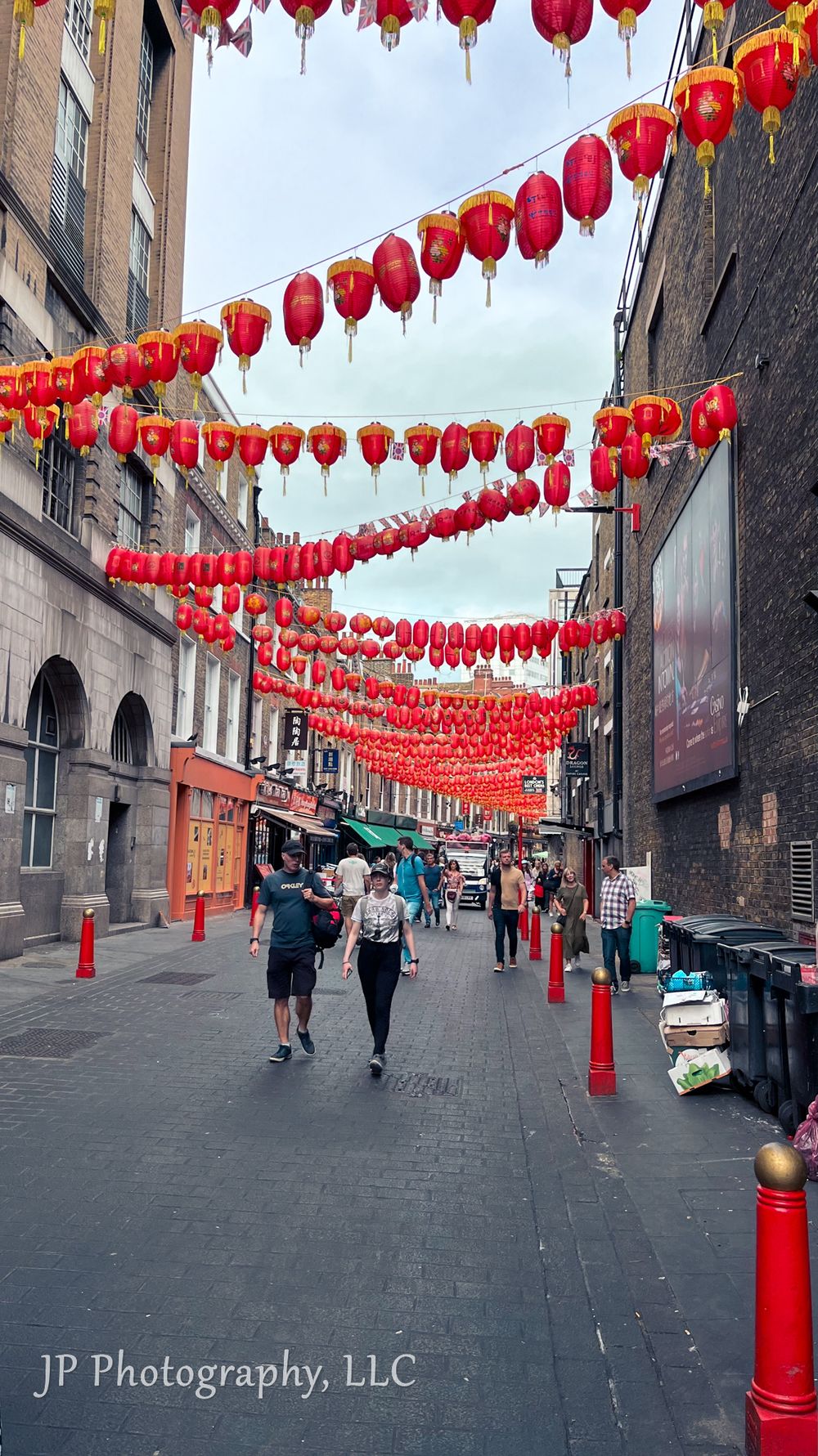 Red Chinese lanterns hang in rows above a narrow urban street with pedestrians walking beneath them.