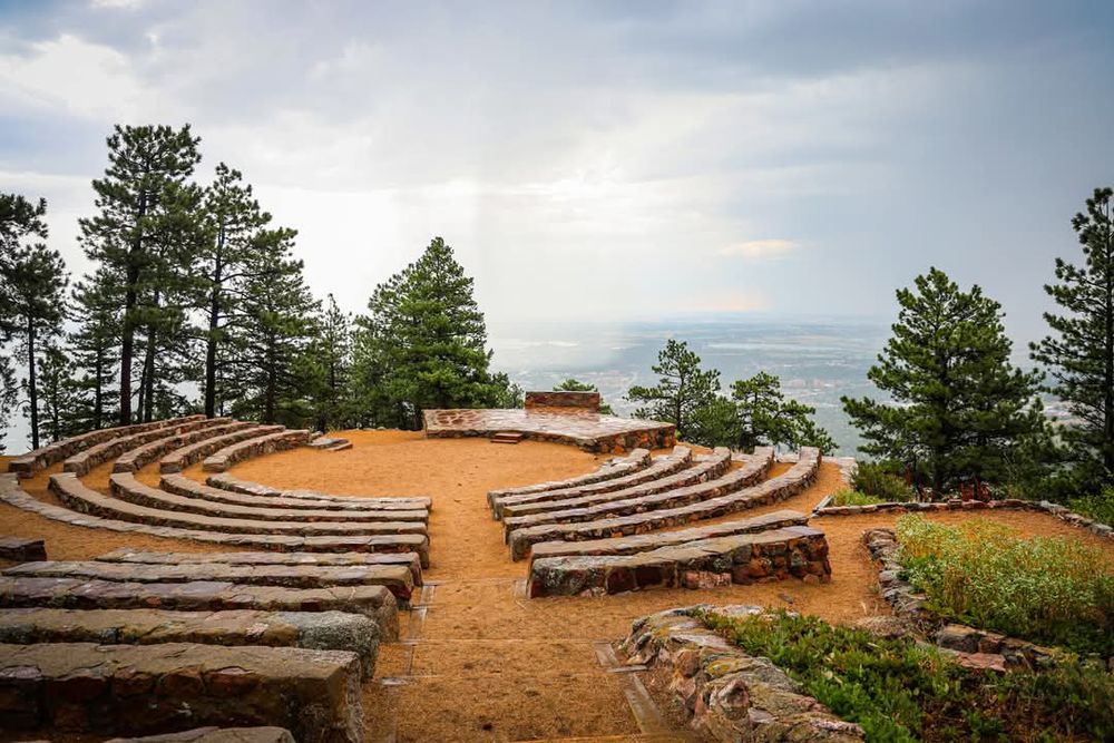 Panoramic view of a rustic outdoor amphitheater with curved stone seating surrounded by pine trees.