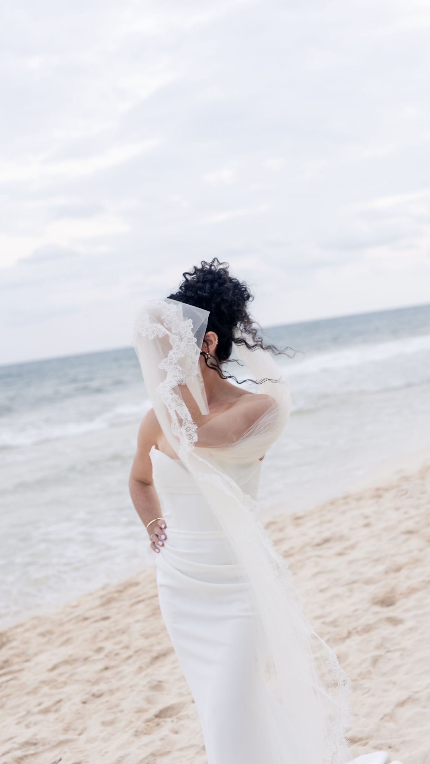 Bride in white wedding dress and veil standing on sandy beach with ocean waves in background on cloudy day.