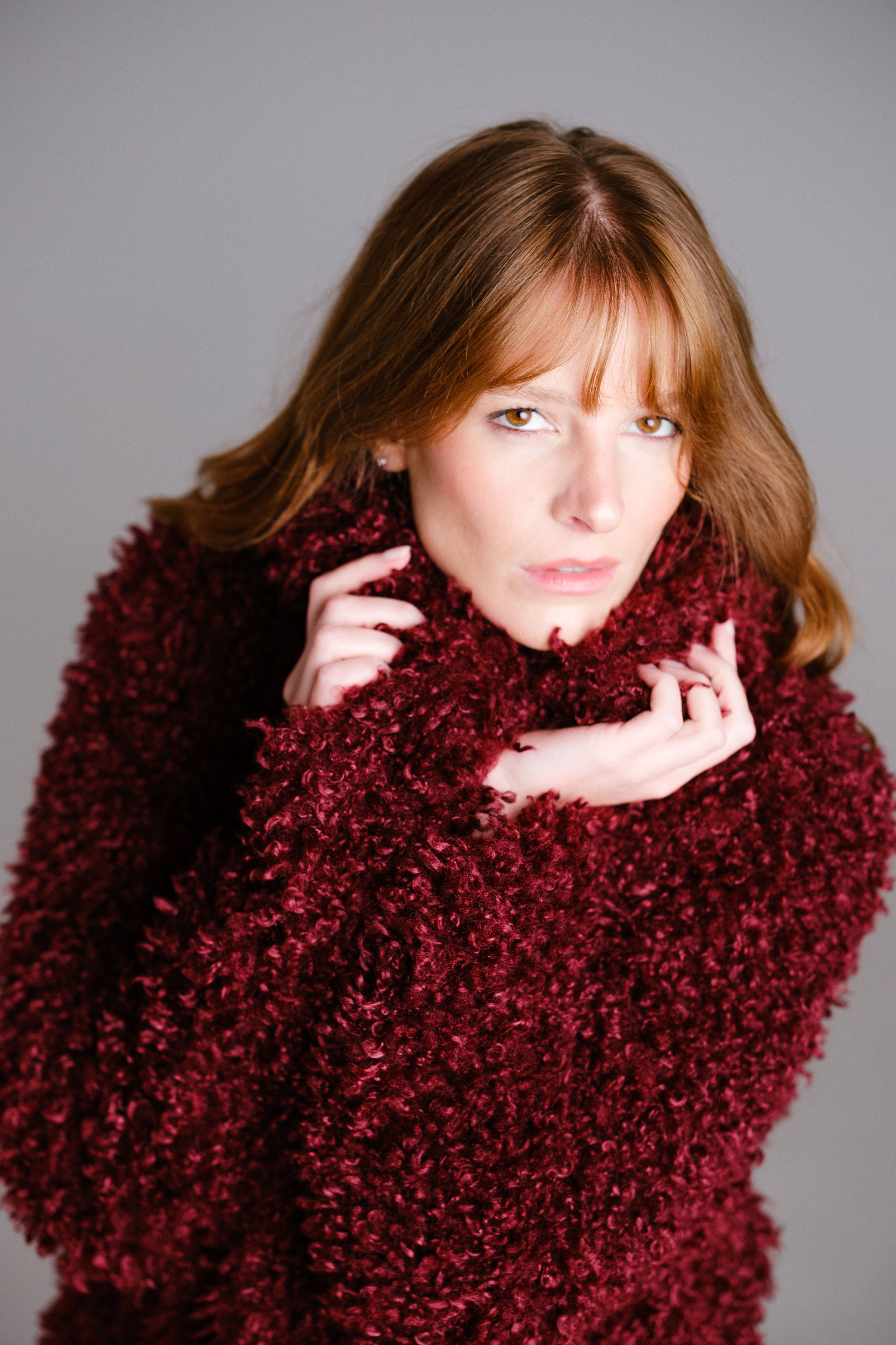 Model in burgundy fuzzy coat poses playfully against grey backdrop in fashion photoshoot series.