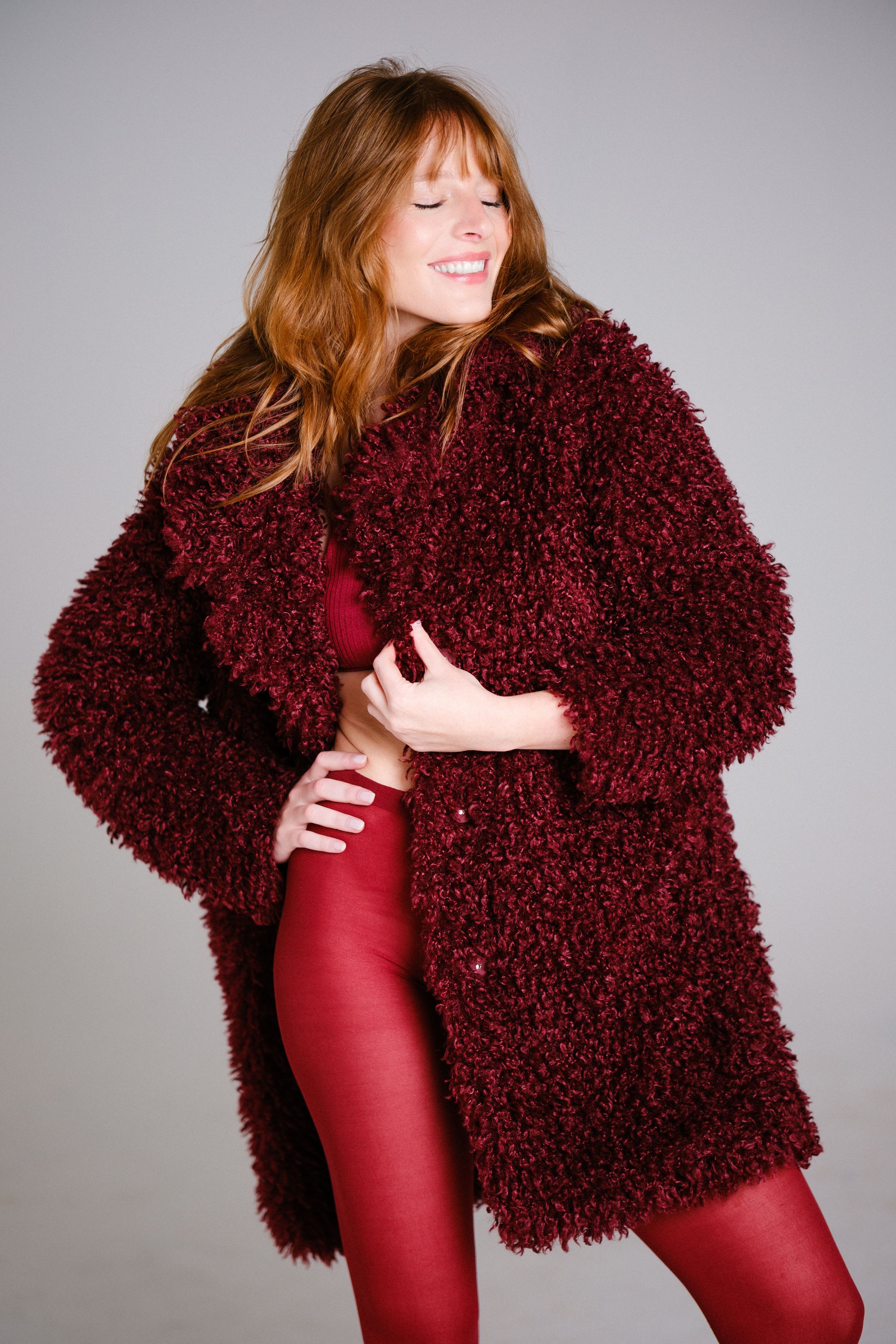Fashion model in monochromatic burgundy outfit with fluffy coat poses expressively.