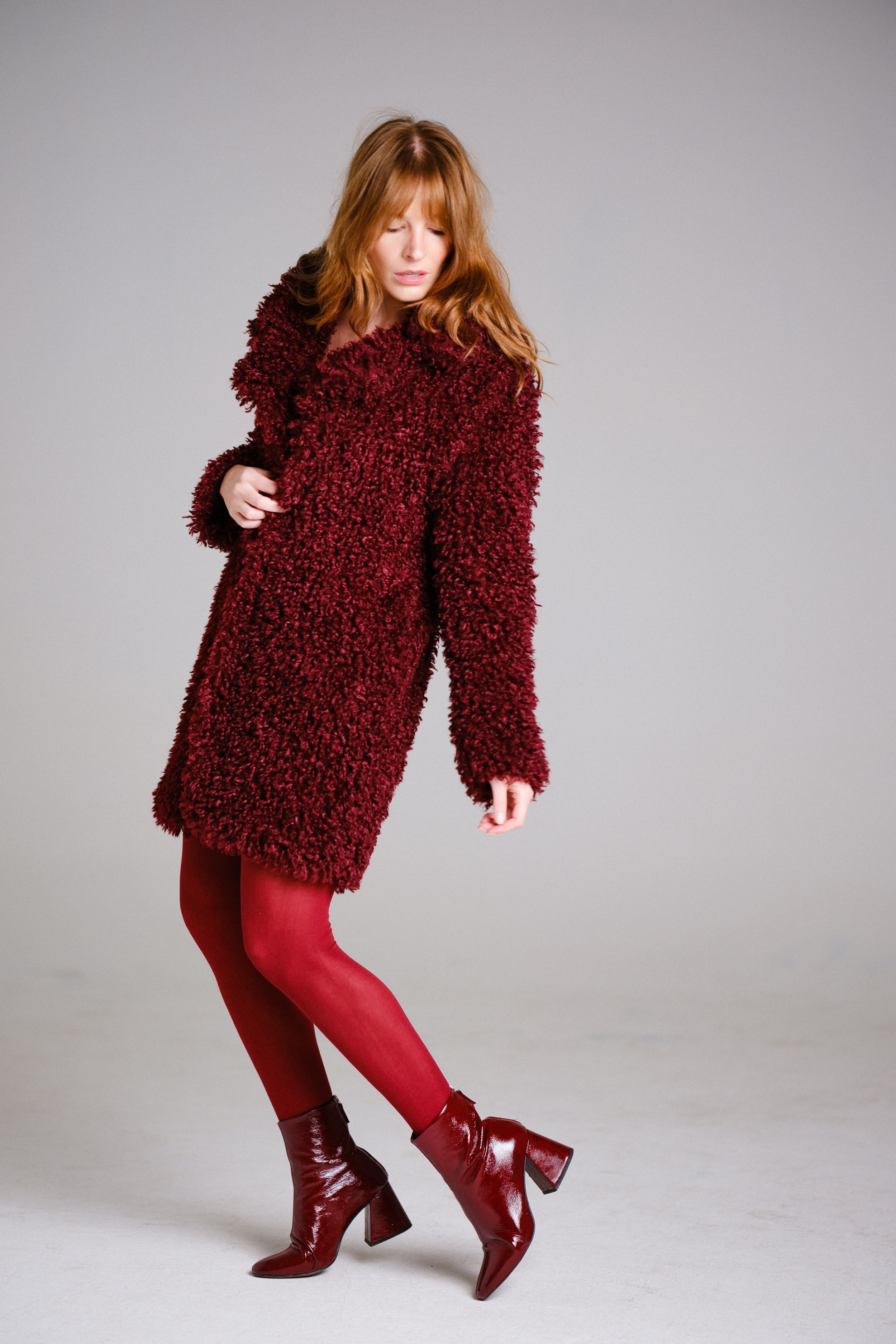 Model showcases burgundy winter look with fluffy coat and matching boots against grey backdrop.