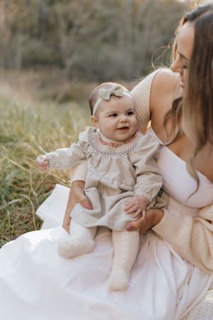 Baby in smocked dress sits with parent in a sunlit field.