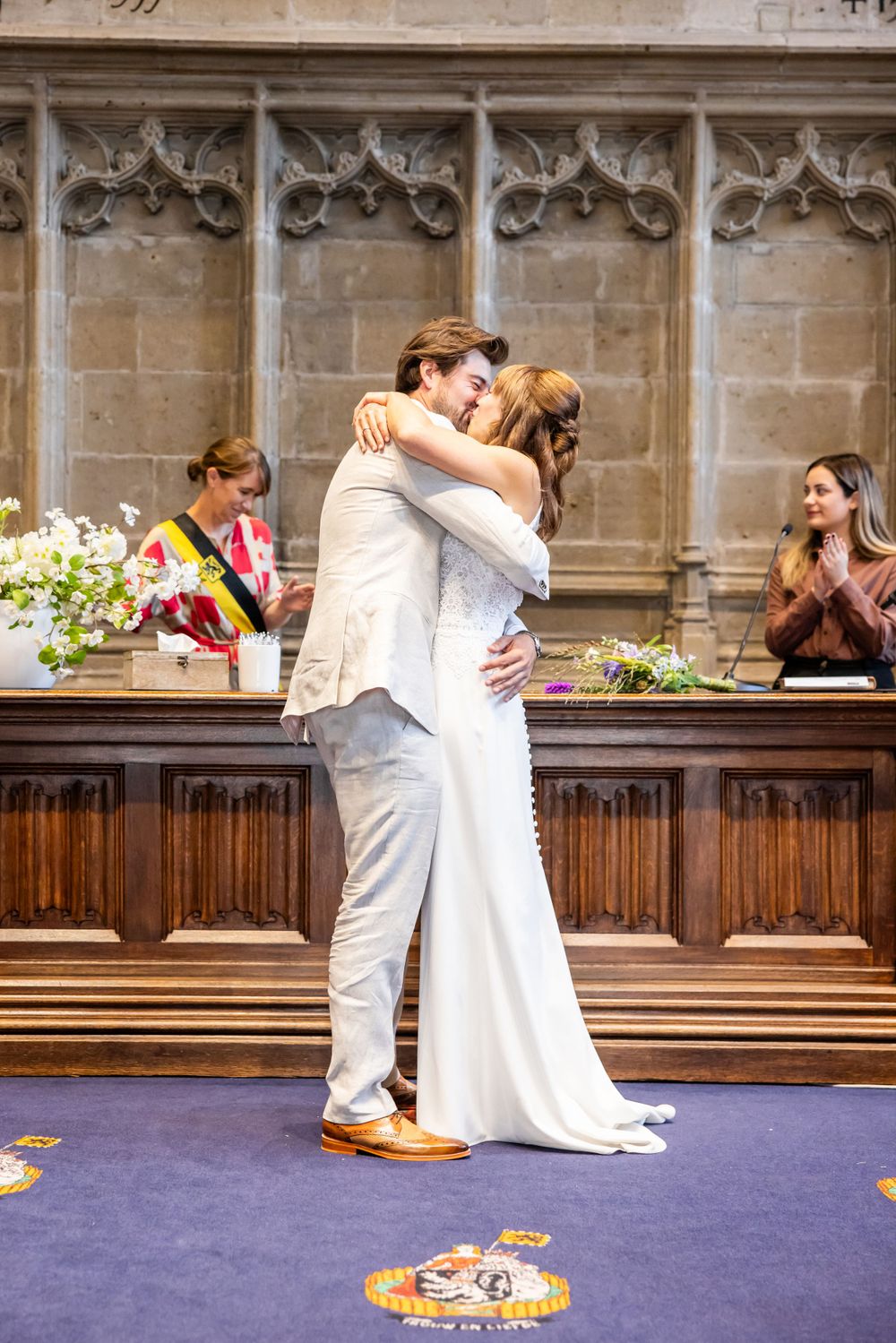 A romantic wedding ceremony in a gothic chapel with ornate stone walls and wooden paneling, on a purple carpeted aisle.