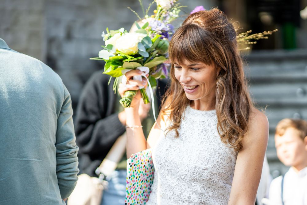 Radiant bride in white lace dress holds bouquet while smiling during outdoor wedding celebration.