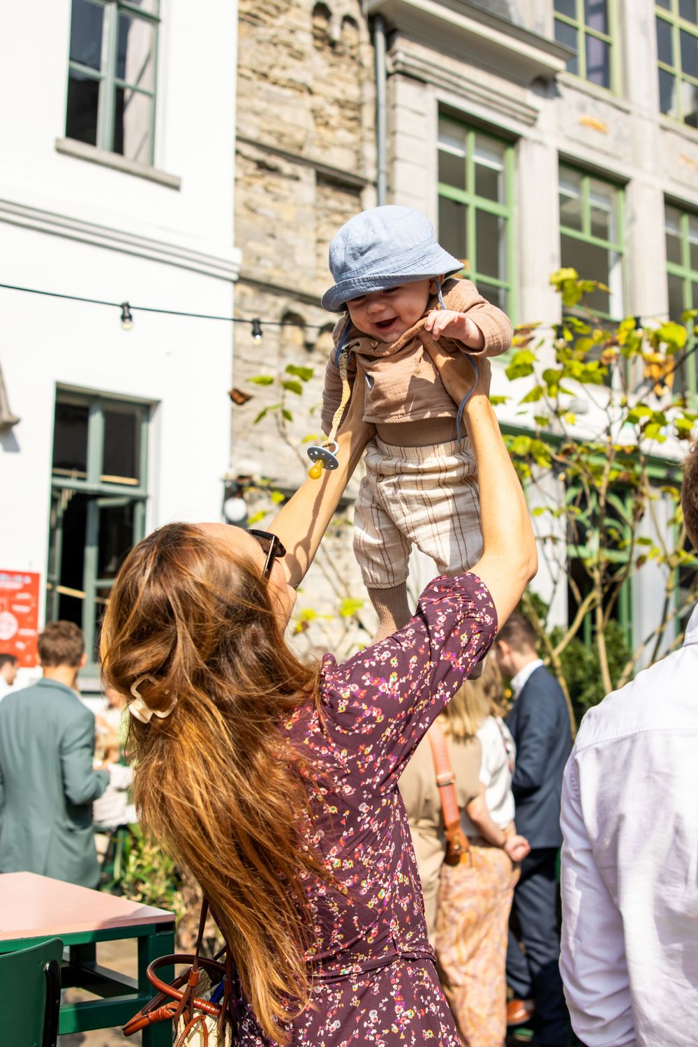 Parent lifting joyful toddler in outdoor cafe setting with stone building facade and greenery in background.
