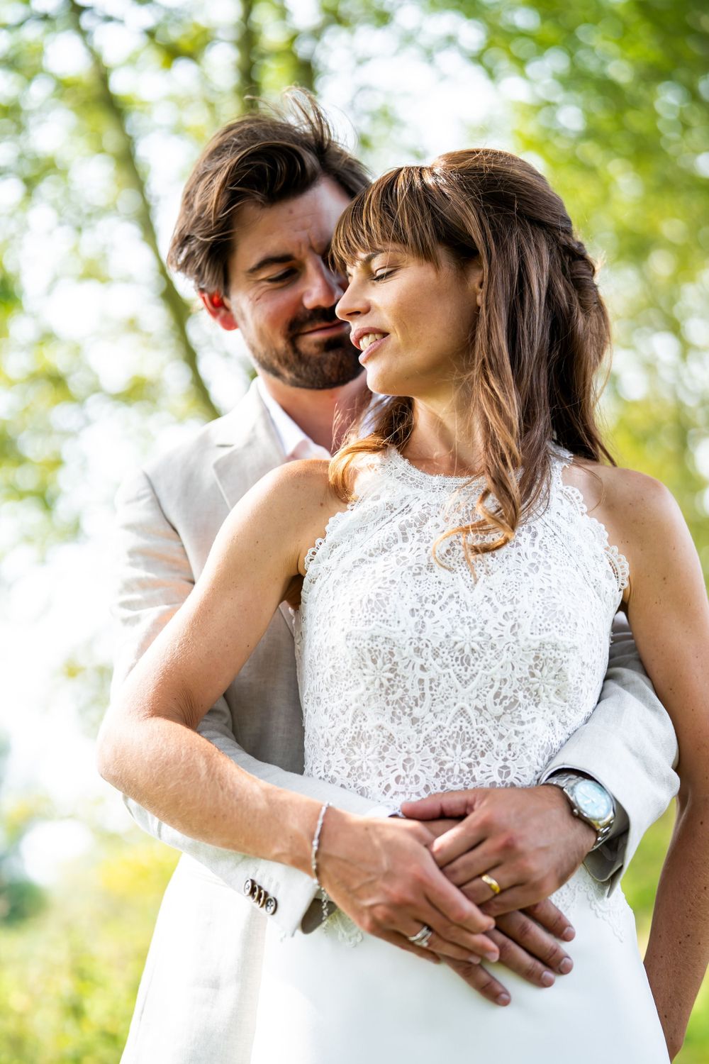 A loving couple embraces outdoors on their wedding day, dressed in white against a soft green bokeh background.