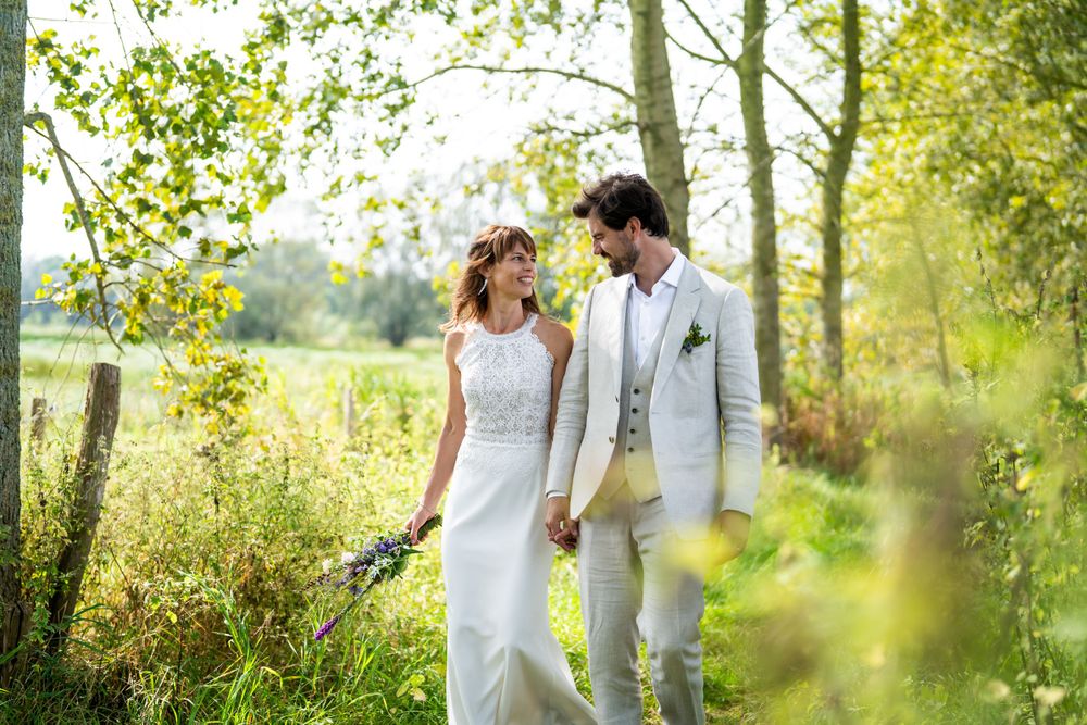 A couple in wedding attire walks together in a sun-dappled meadow surrounded by trees on a bright summer day.