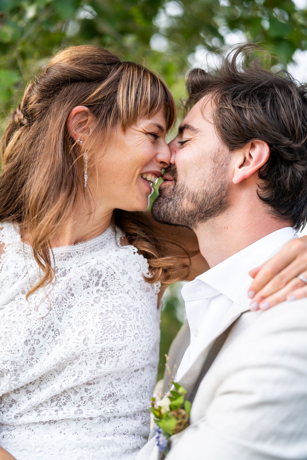 A romantic couple in wedding attire share an intimate moment outdoors, the bride in white lace and groom in light gray.