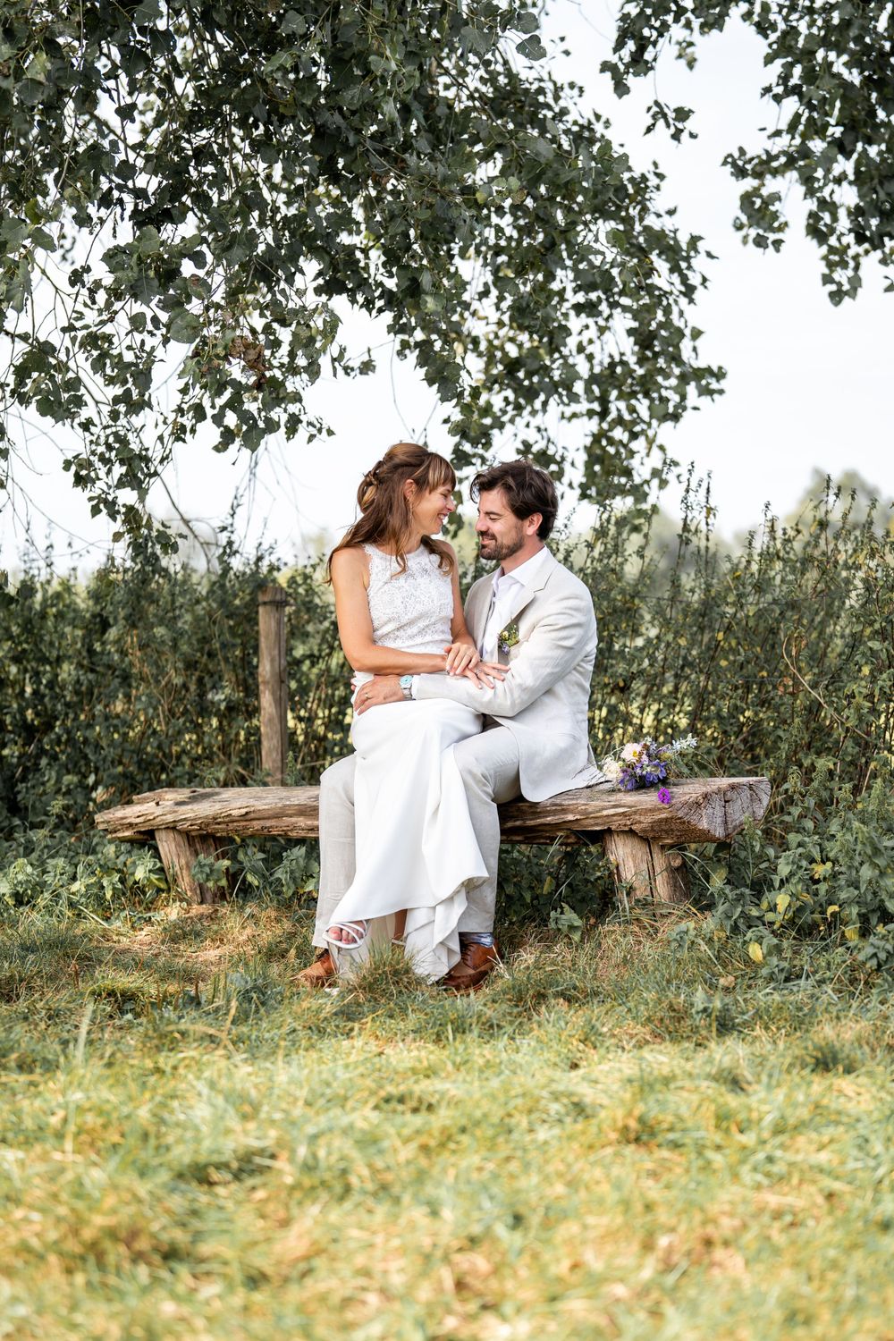 A couple in white wedding attire sits together on a rustic wooden bench surrounded by lush greenery and trees.