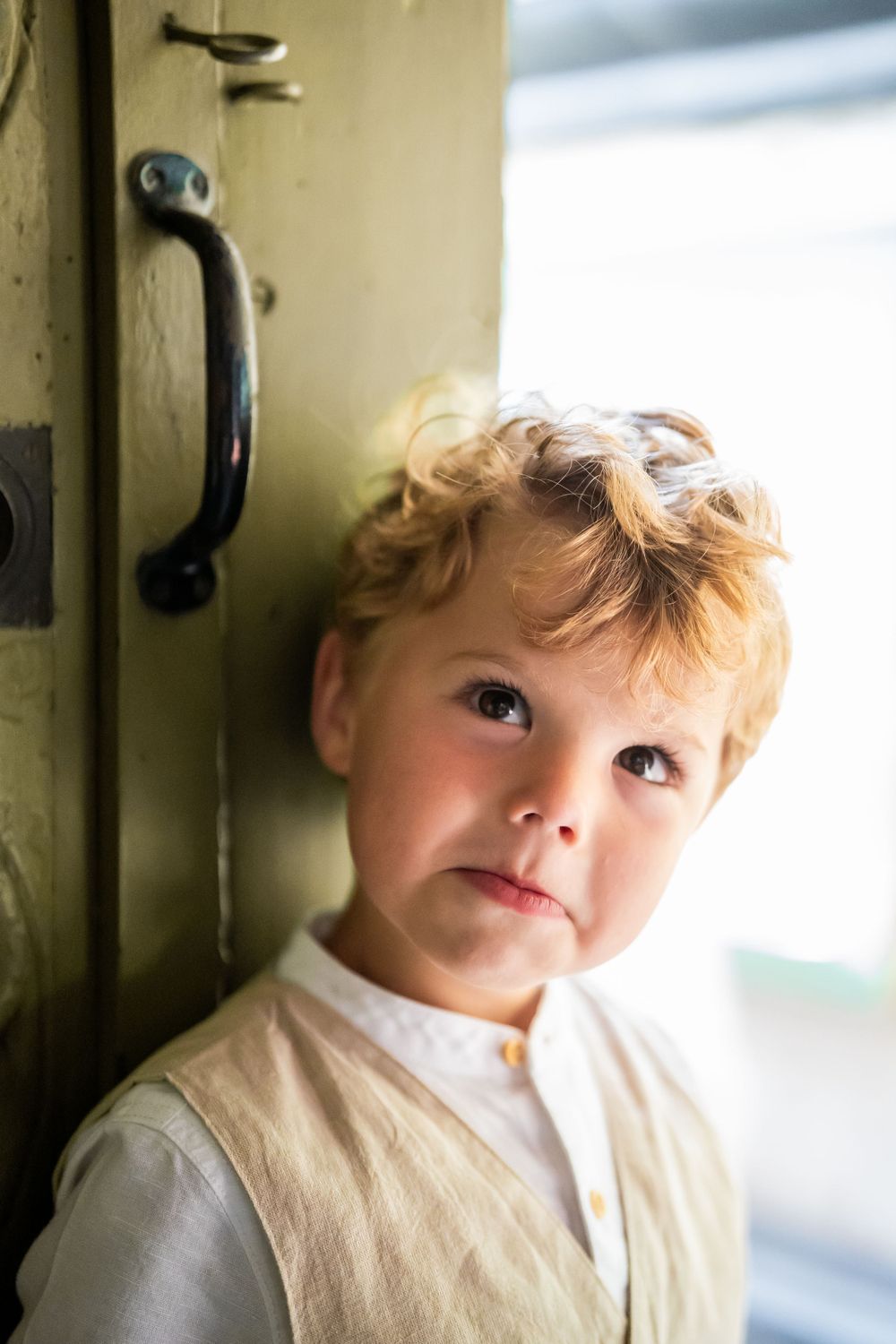 A young child with tousled blonde hair stands near a door handle in soft natural light.