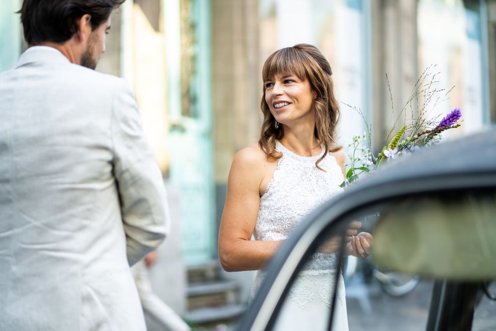 A couple in wedding attire smiles at each other near a car while holding a delicate floral bouquet.