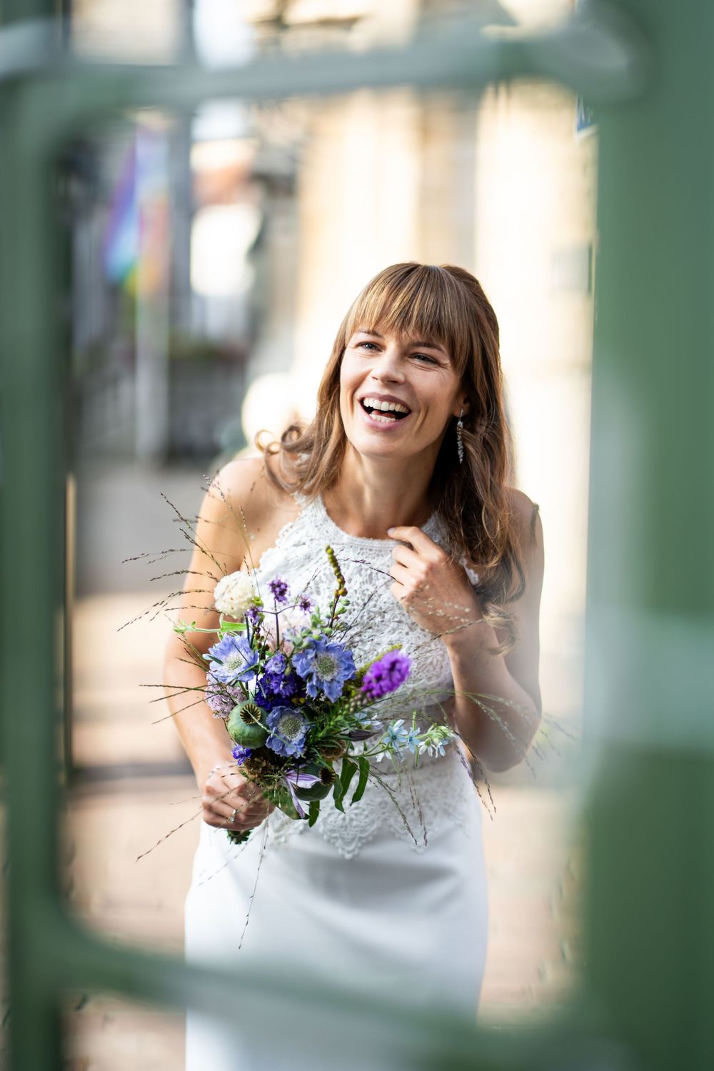 Joyful bride in white dress laughs while holding a vibrant purple and blue wildflower bouquet.
