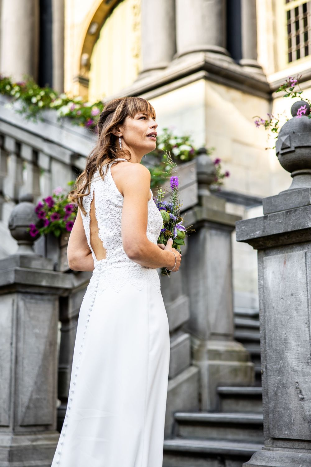 A bride in a white lace wedding dress holds purple flowers while standing on stone steps near ornate architecture.