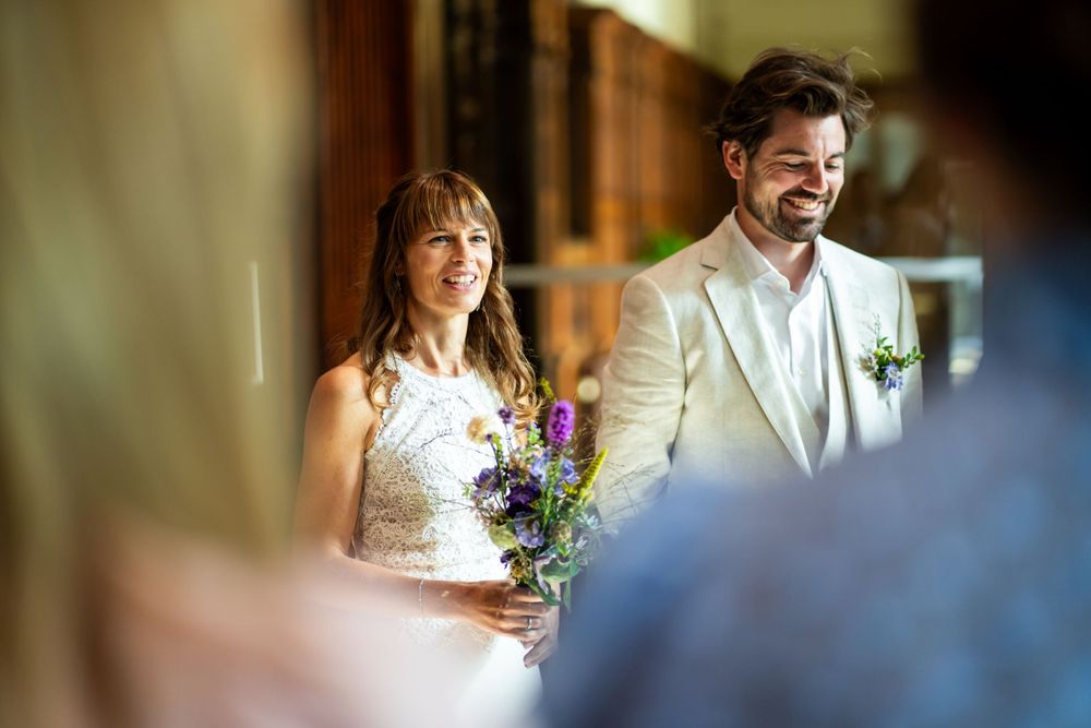 A couple stands together at their wedding ceremony, with the bride holding a purple and white bouquet.