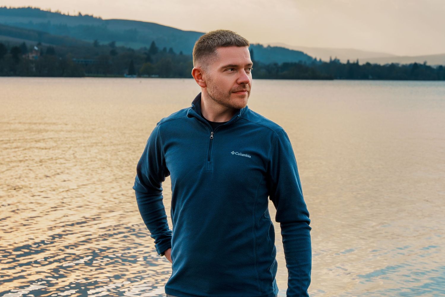 Scotty D, UK freelance photographer, wearing a blue Columbia jacket, standing by a lake with scenic hills