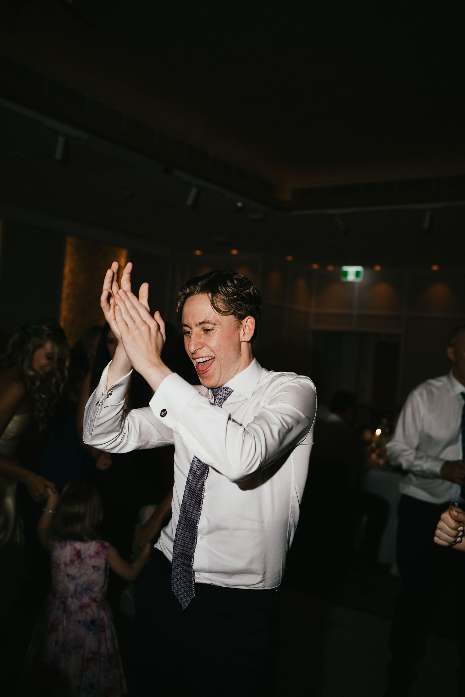A guest in formal attire claps enthusiastically while dancing at a wedding reception.