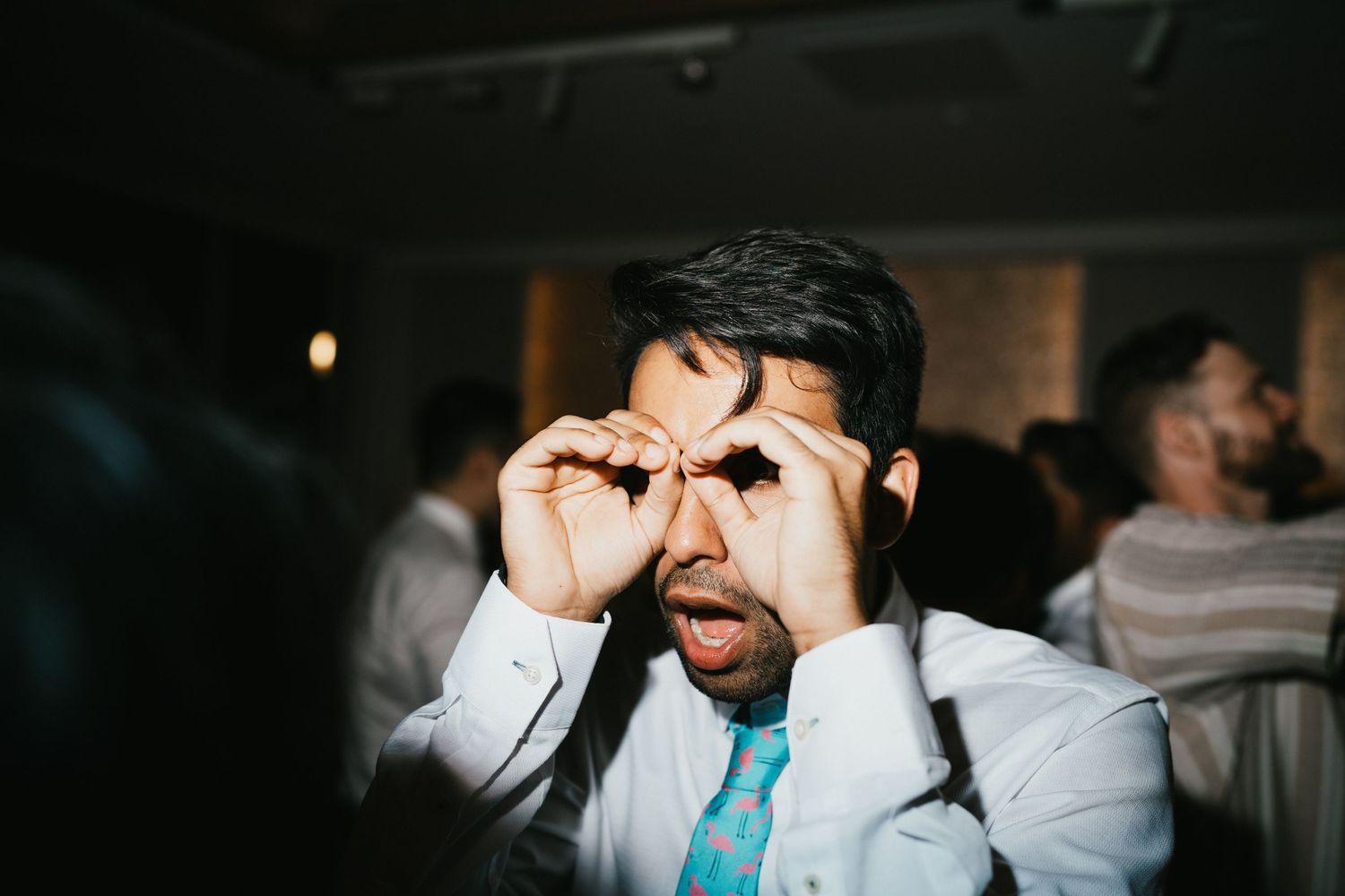 A guest in a white shirt and turquoise tie makes a playful gesture at a party.