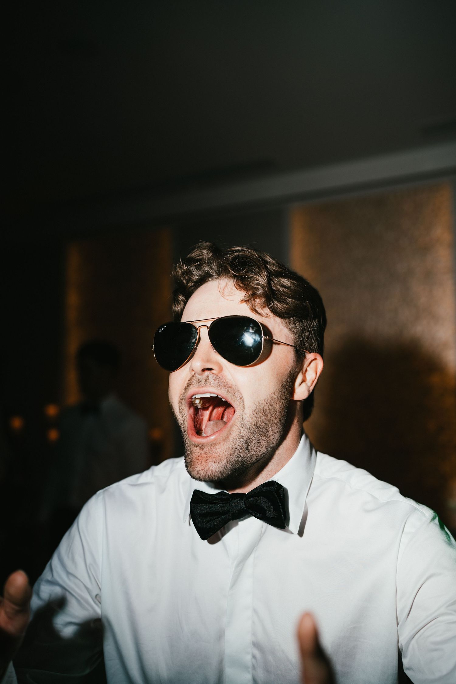 A party guest wearing sunglasses and a bowtie sings enthusiastically at a celebration.