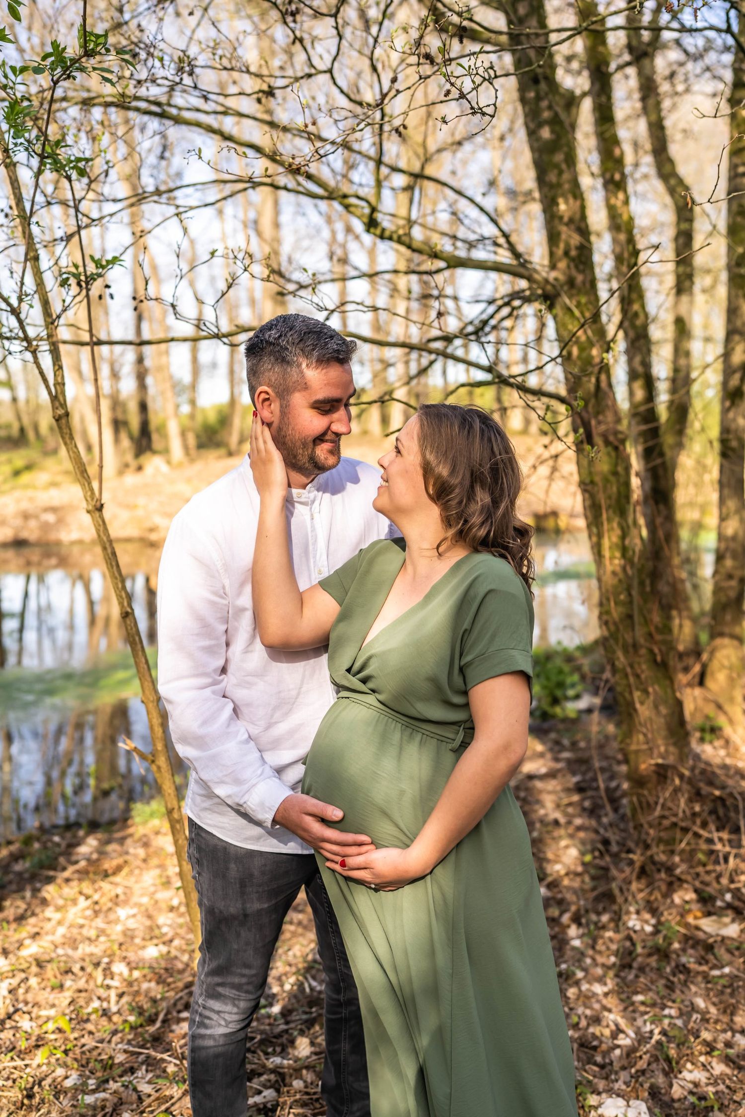 An expecting couple shares an intimate moment in a wooded area with water in the background.