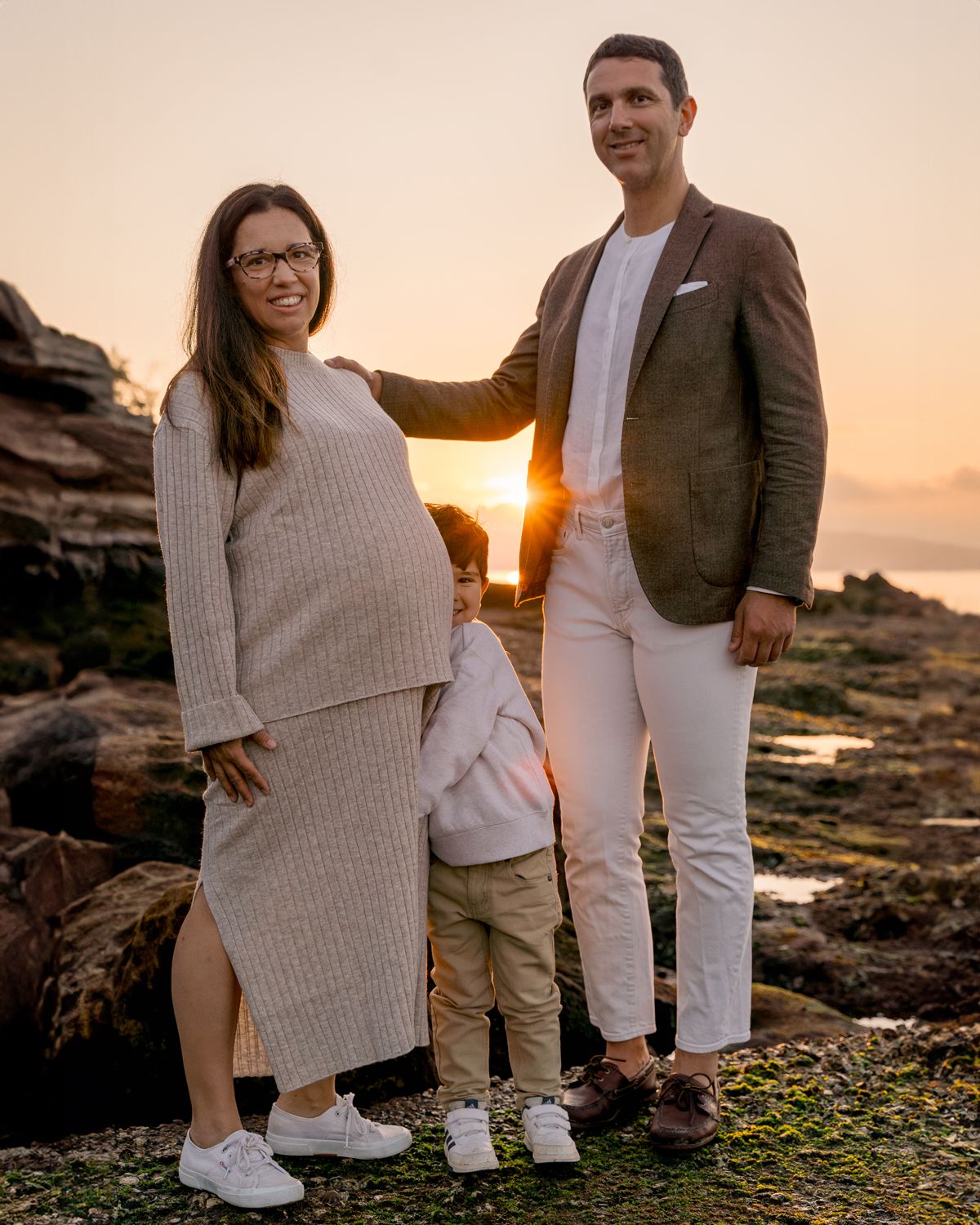 Mom and dad smiling while their son hides playfully behind mom, with the sun peeking through a sunrise family photoshoot