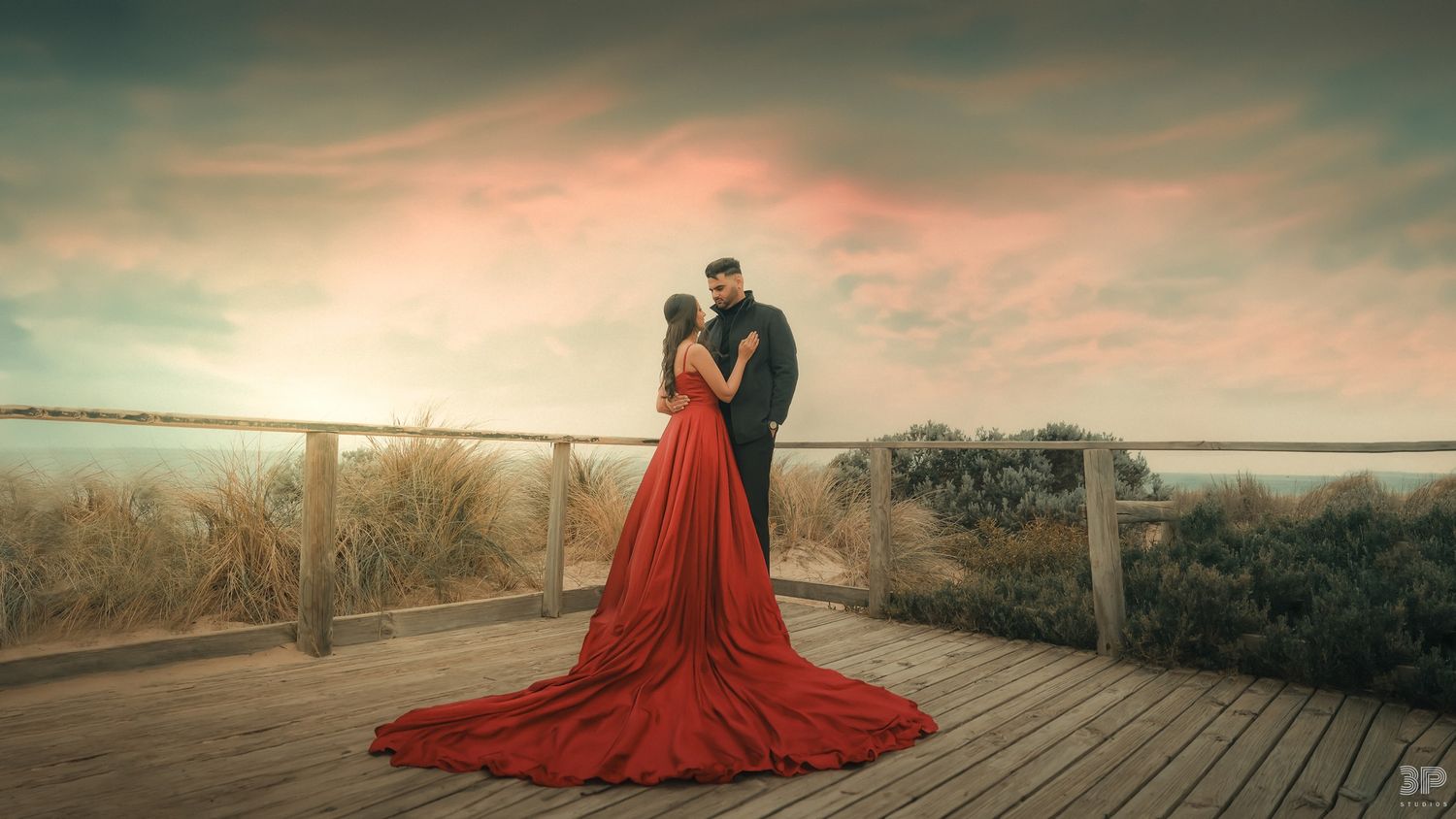 Dramatic sunset photo of couple on wooden deck with flowing red gown.