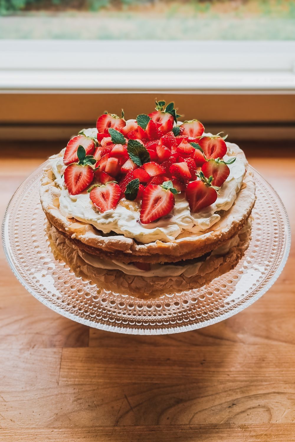 A layered cream cake topped with fresh strawberries on a glass cake stand.