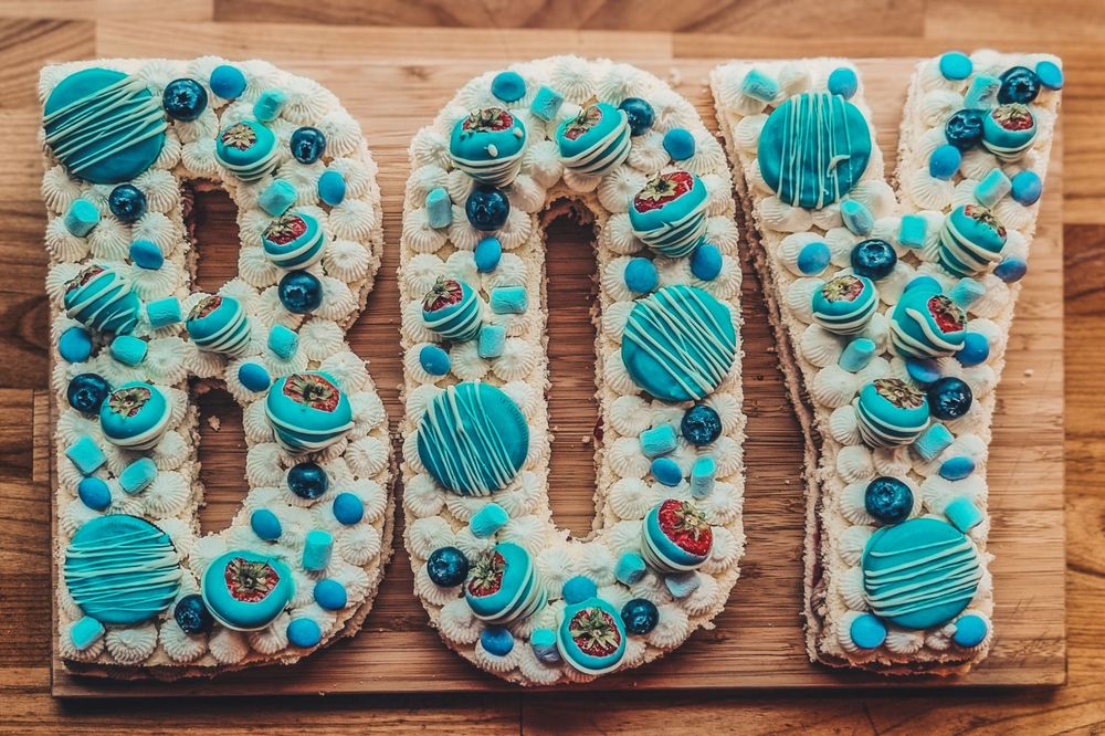 Letter-shaped cream cakes spelling 'BOY' decorated with blue macarons and sprinkles.