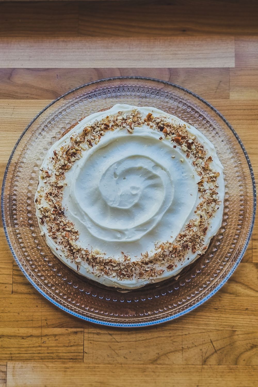 A cream-frosted cake with a swirled top and crushed nuts around the edges on a glass plate.