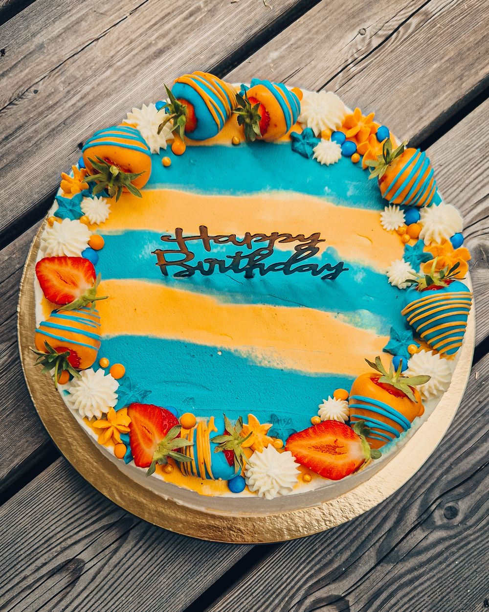 Blue and orange striped birthday cake decorated with chocolate covered strawberries and swirl candies sits on wooden deck.