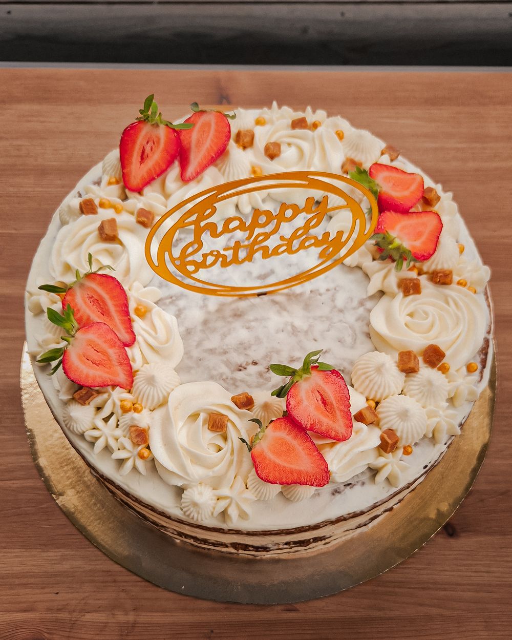 Classic birthday cake topped with whipped cream, fresh strawberries, and caramel writing on a wooden table.