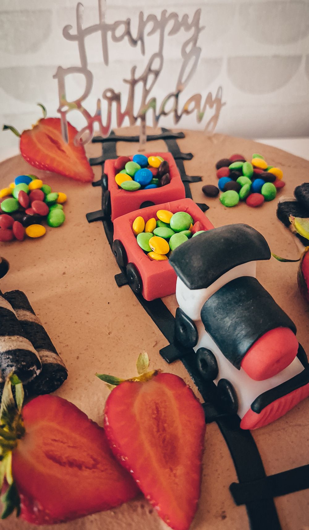 A chocolate birthday cake decorated with train carriages filled with M&Ms and fresh strawberries.