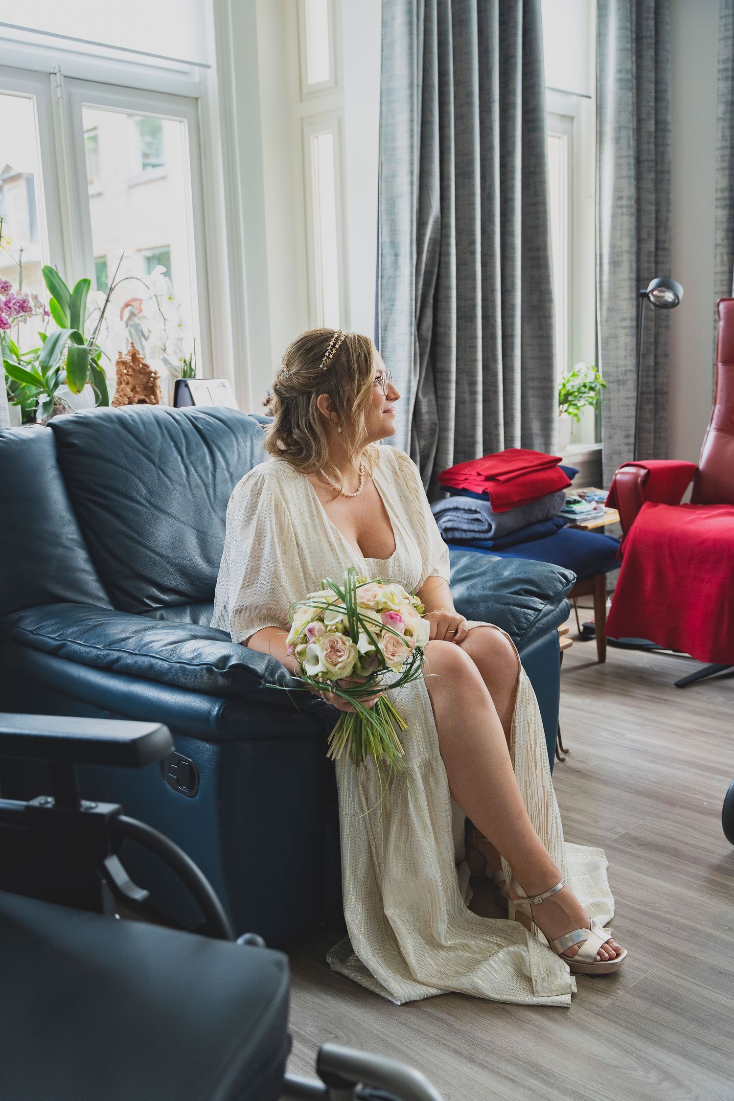 A bride in a vintage-style dress sits on a blue couch holding a bouquet near a bright window.