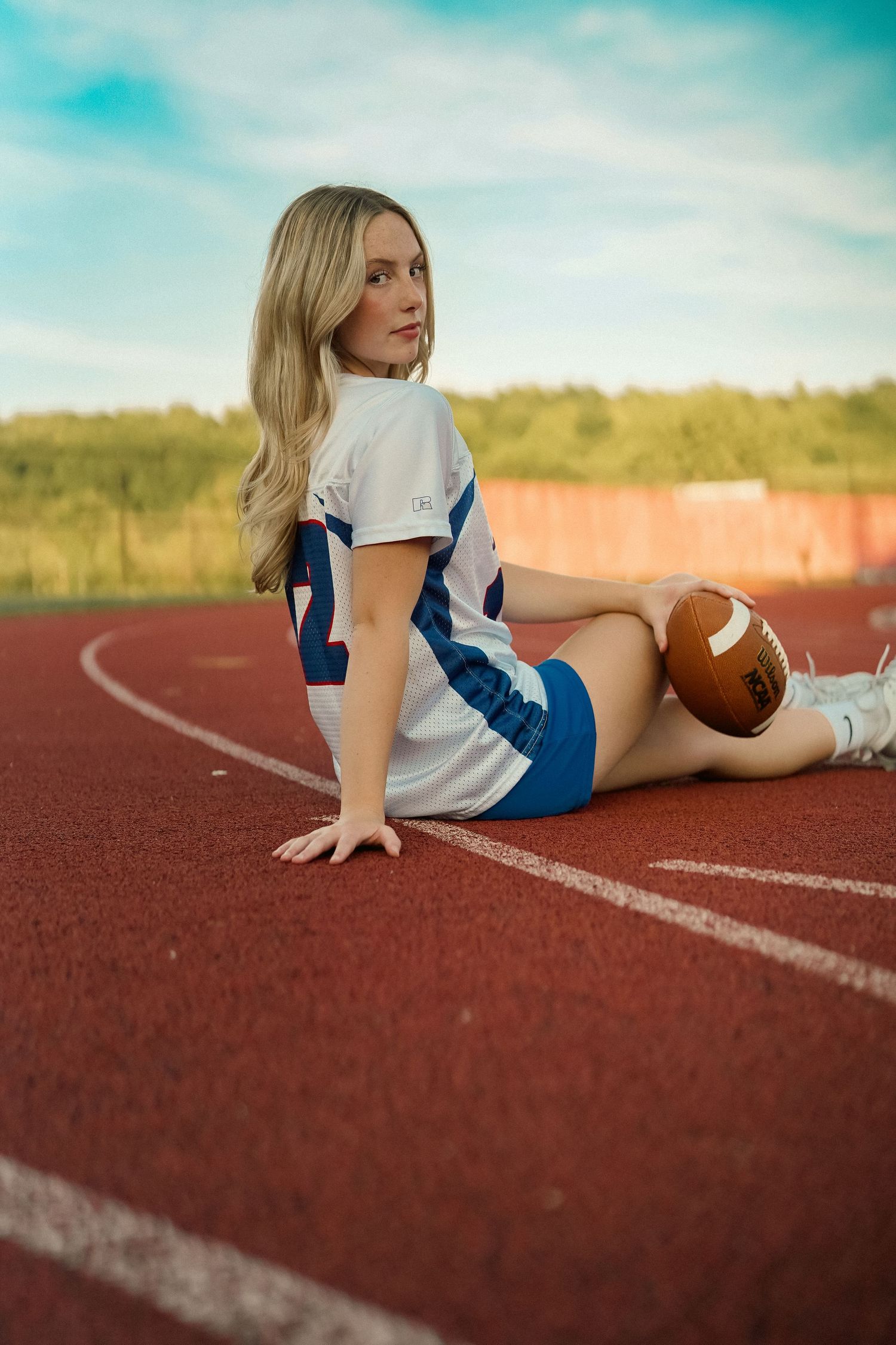 Athletic model in football jersey poses with ball on red running track.