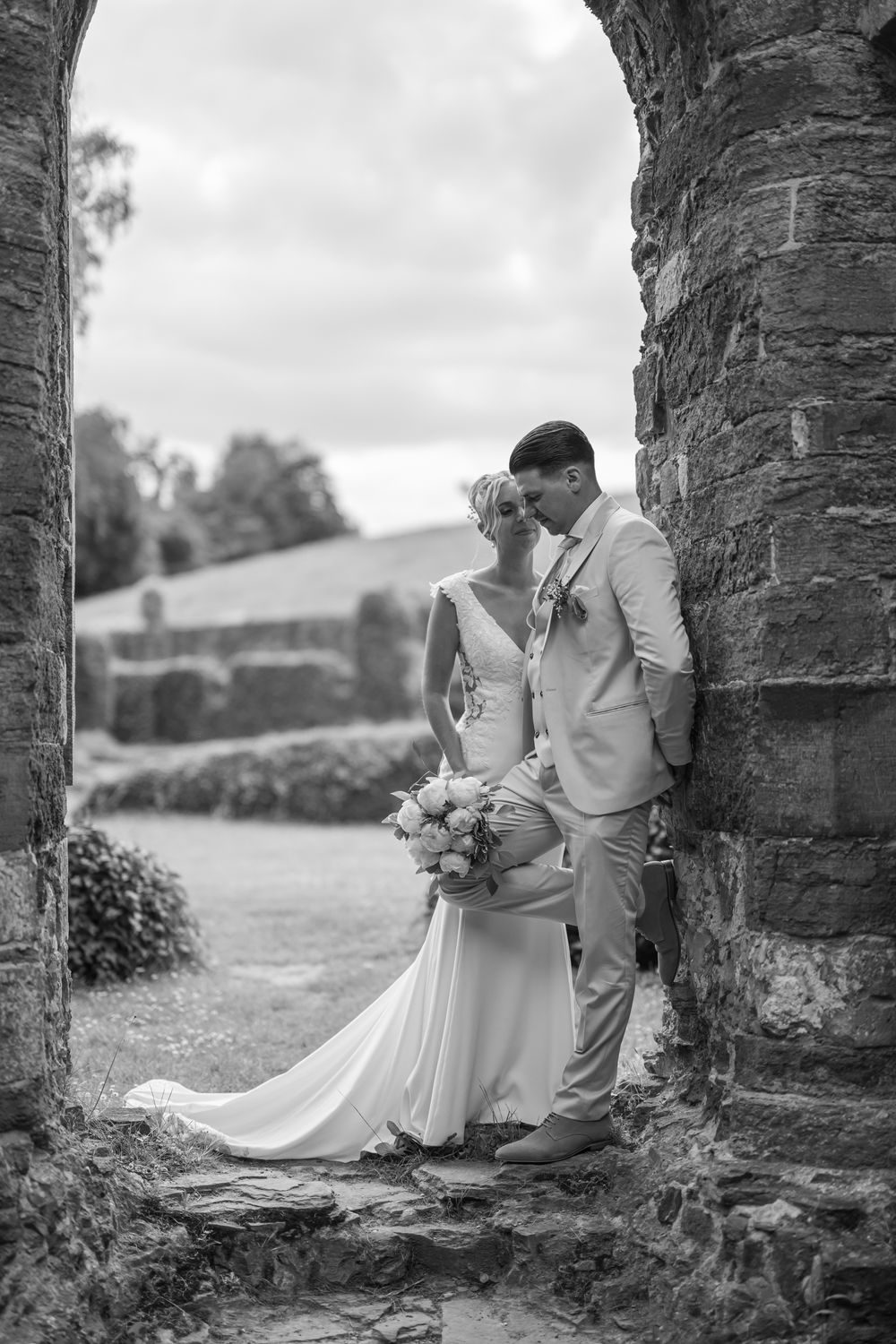 Black and white romantic portrait of newlyweds framed by stone archway in countryside setting.