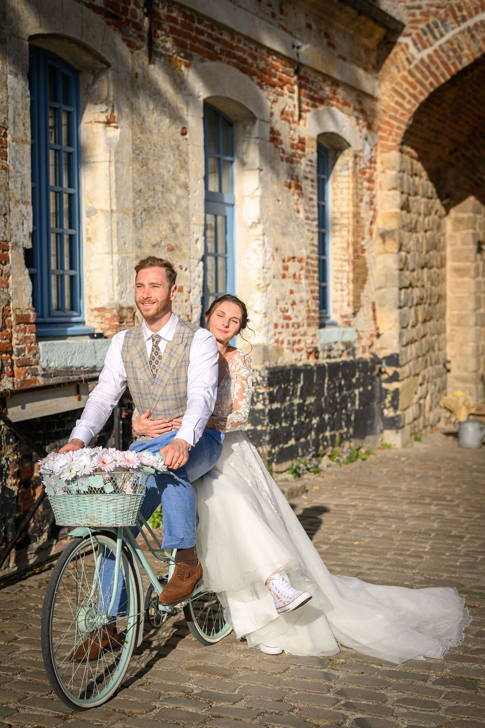Romantic wedding couple poses with vintage turquoise bicycle against rustic brick archway and cobblestone path.