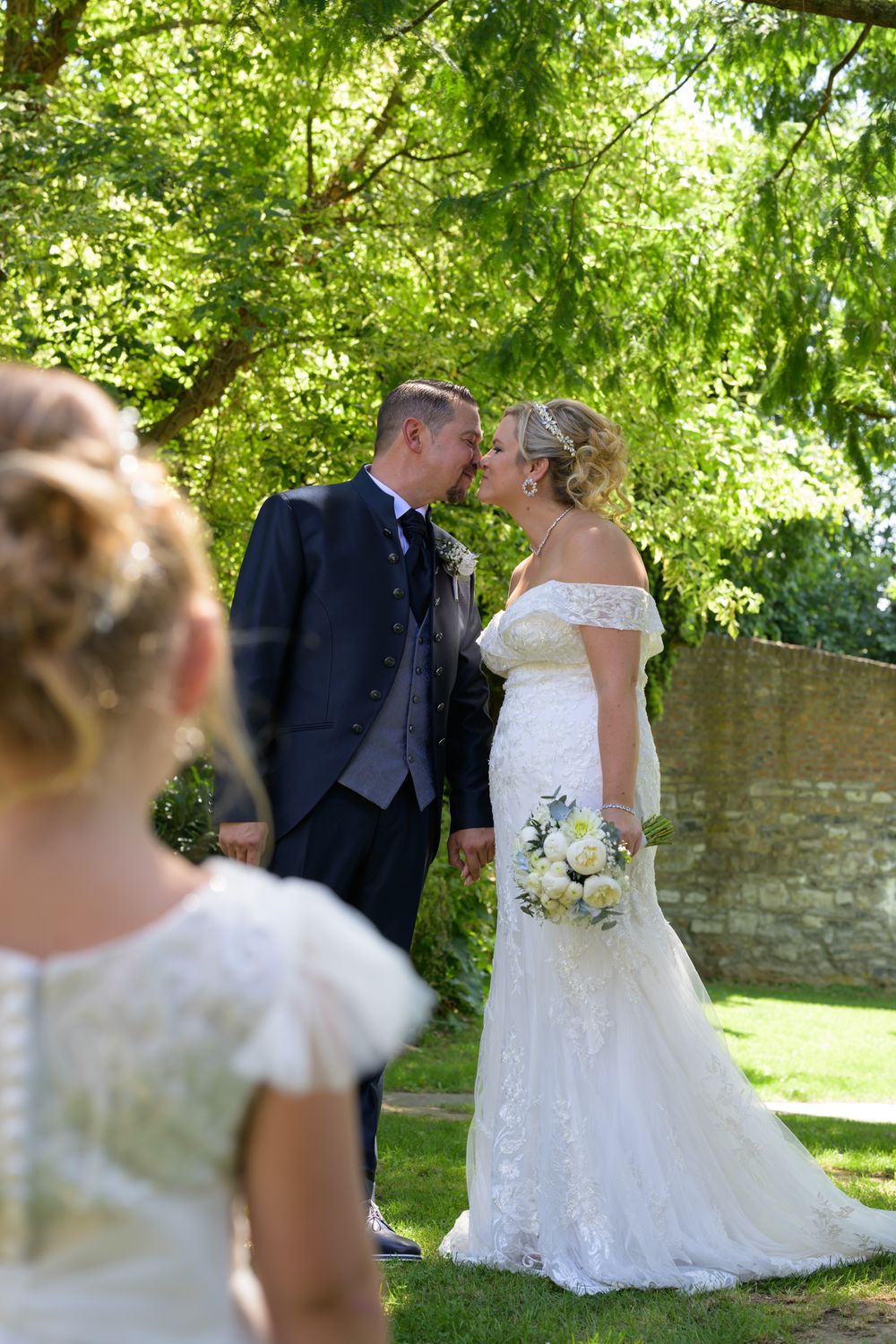 Romantic newlyweds share tender moments under lush green trees near a stone wall.