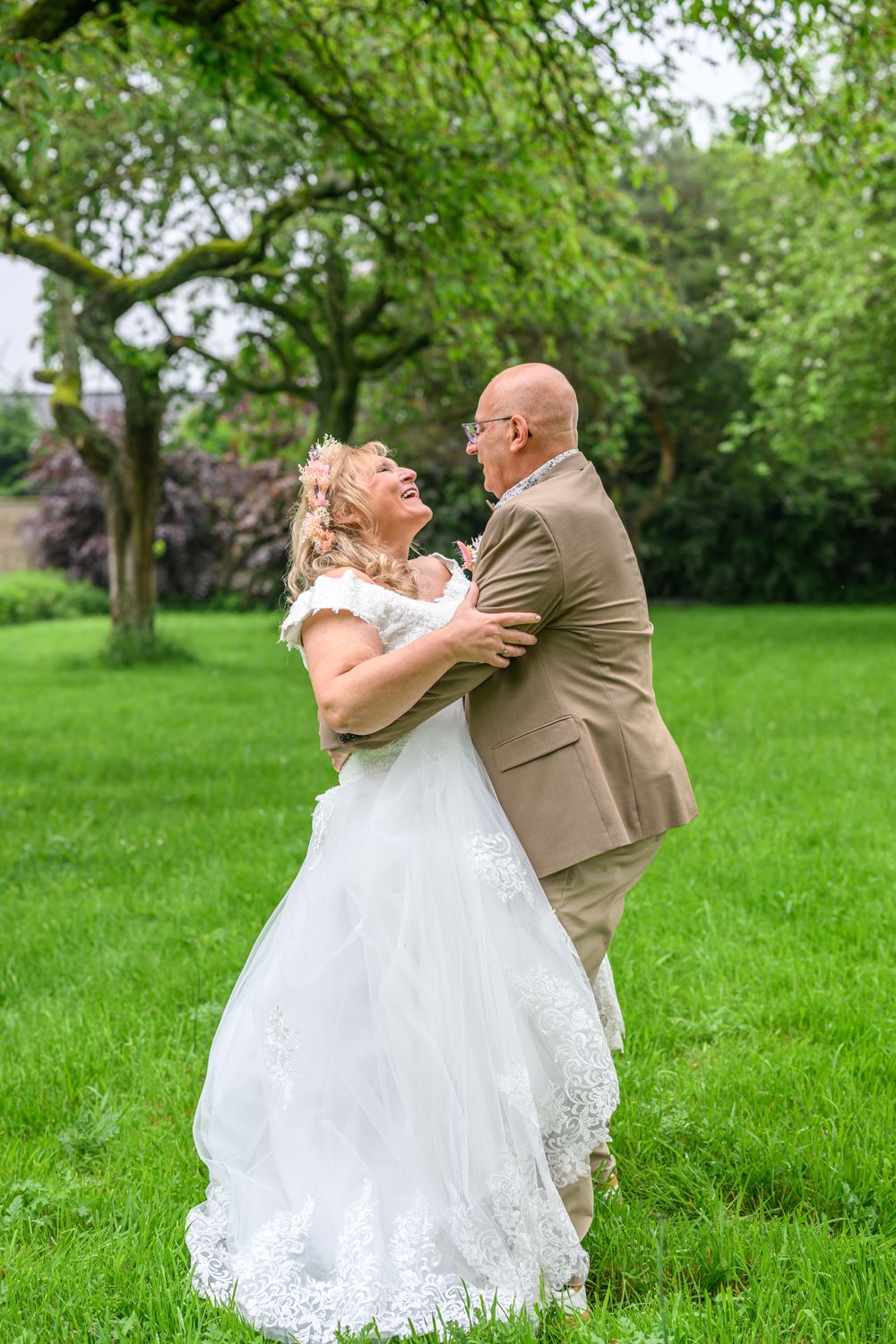 Wedding couple dances and laughs together on a lush green lawn under tree canopy.