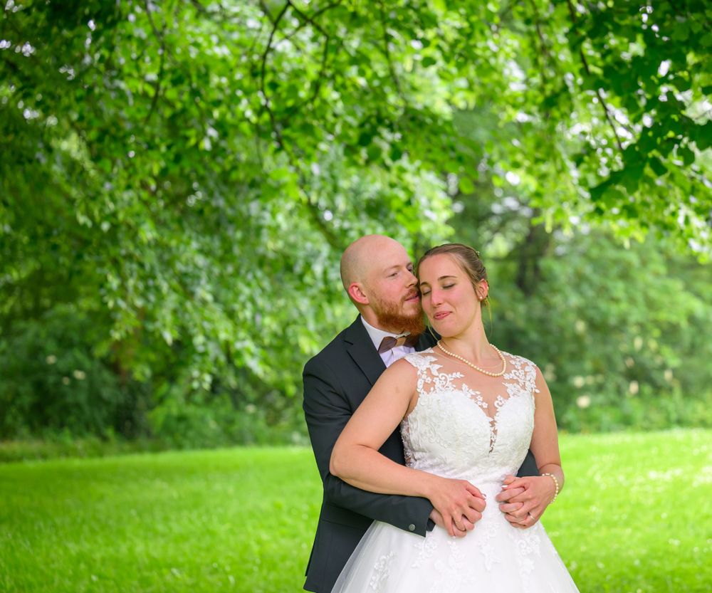 Romantic wedding couple embraces under green summer foliage in natural outdoor setting.