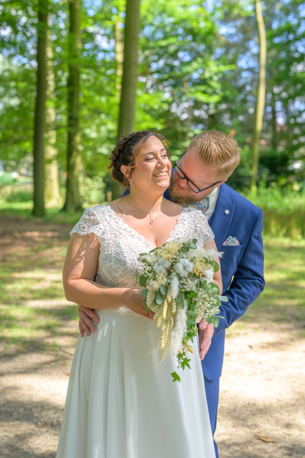 Joyful wedding couple shares intimate moments in a sunlit wooded area with cascading floral bouquet.