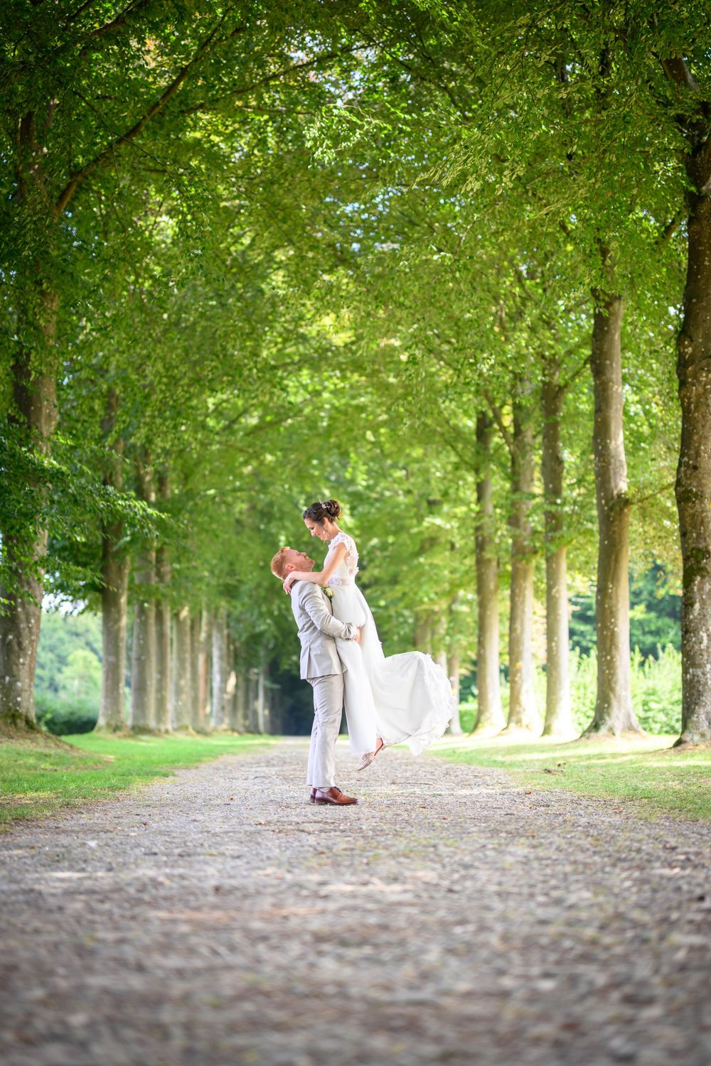 A romantic tree-lined path in a park shows a couple walking together in the distance on a sunny summer day.