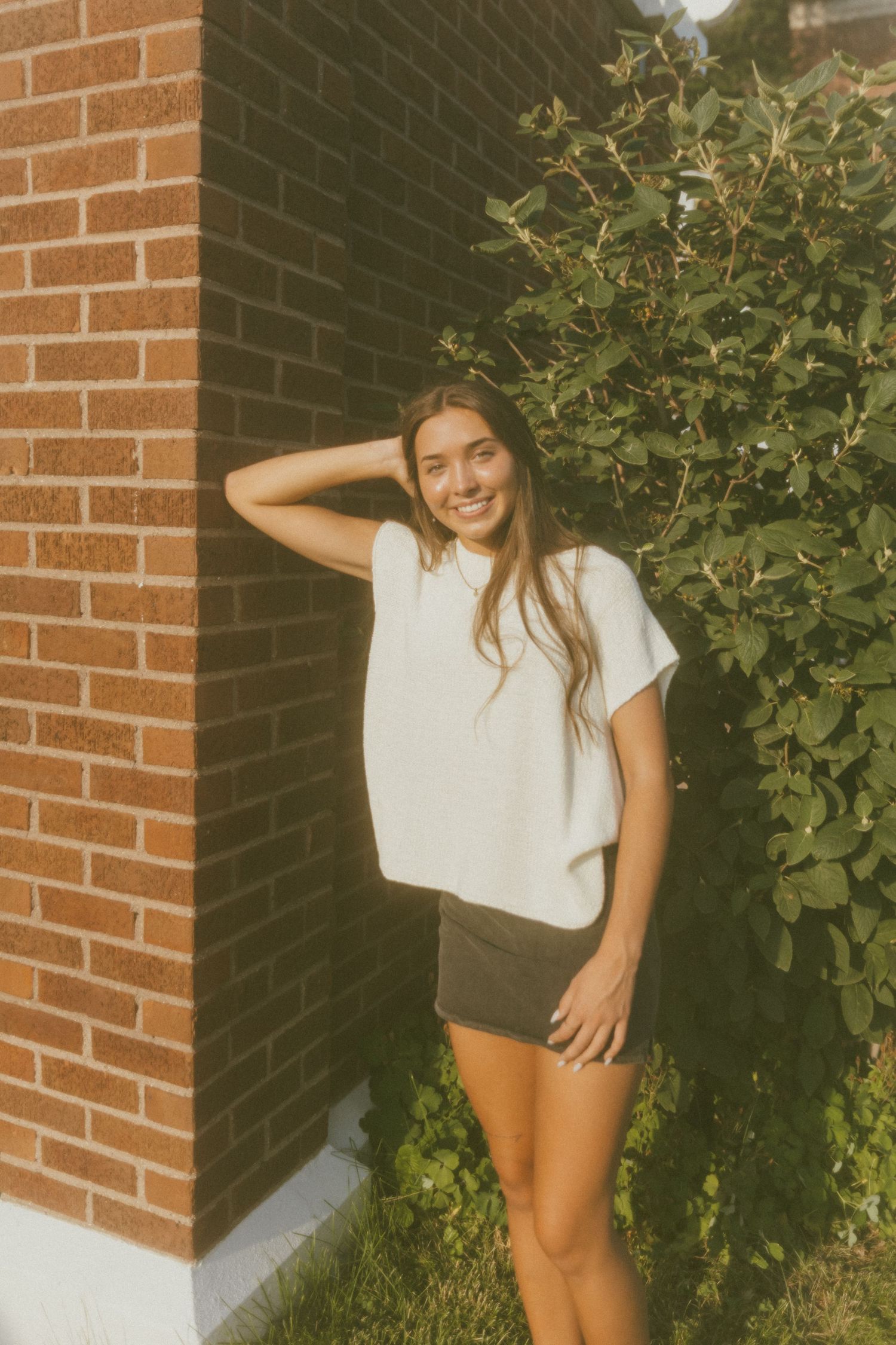 Smiling young woman in white tee and dark shorts posing by a brick wall with green bushes outdoors.