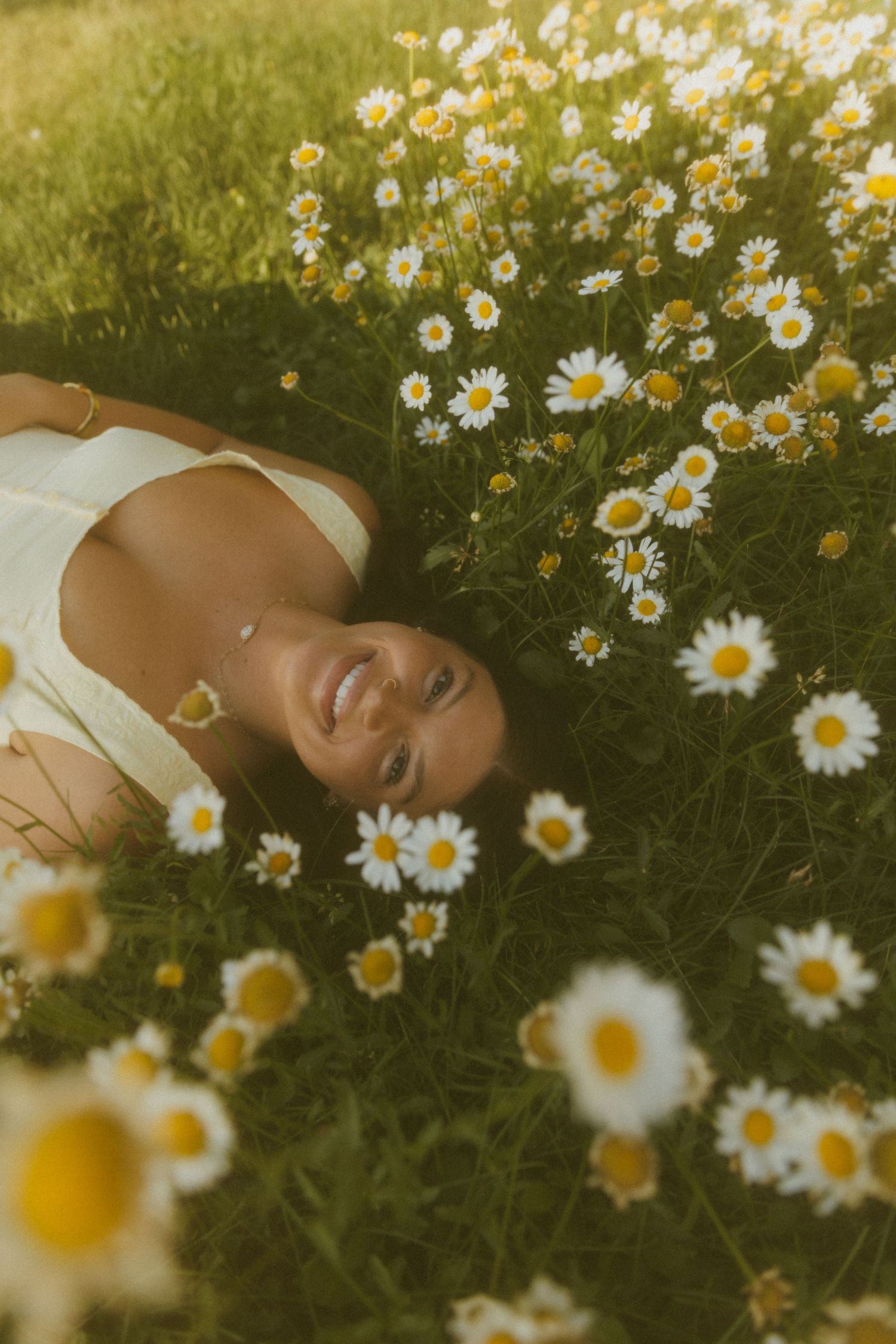 A smiling woman lying in a field of white daisies, bathed in warm golden sunlight, wearing a white top.
