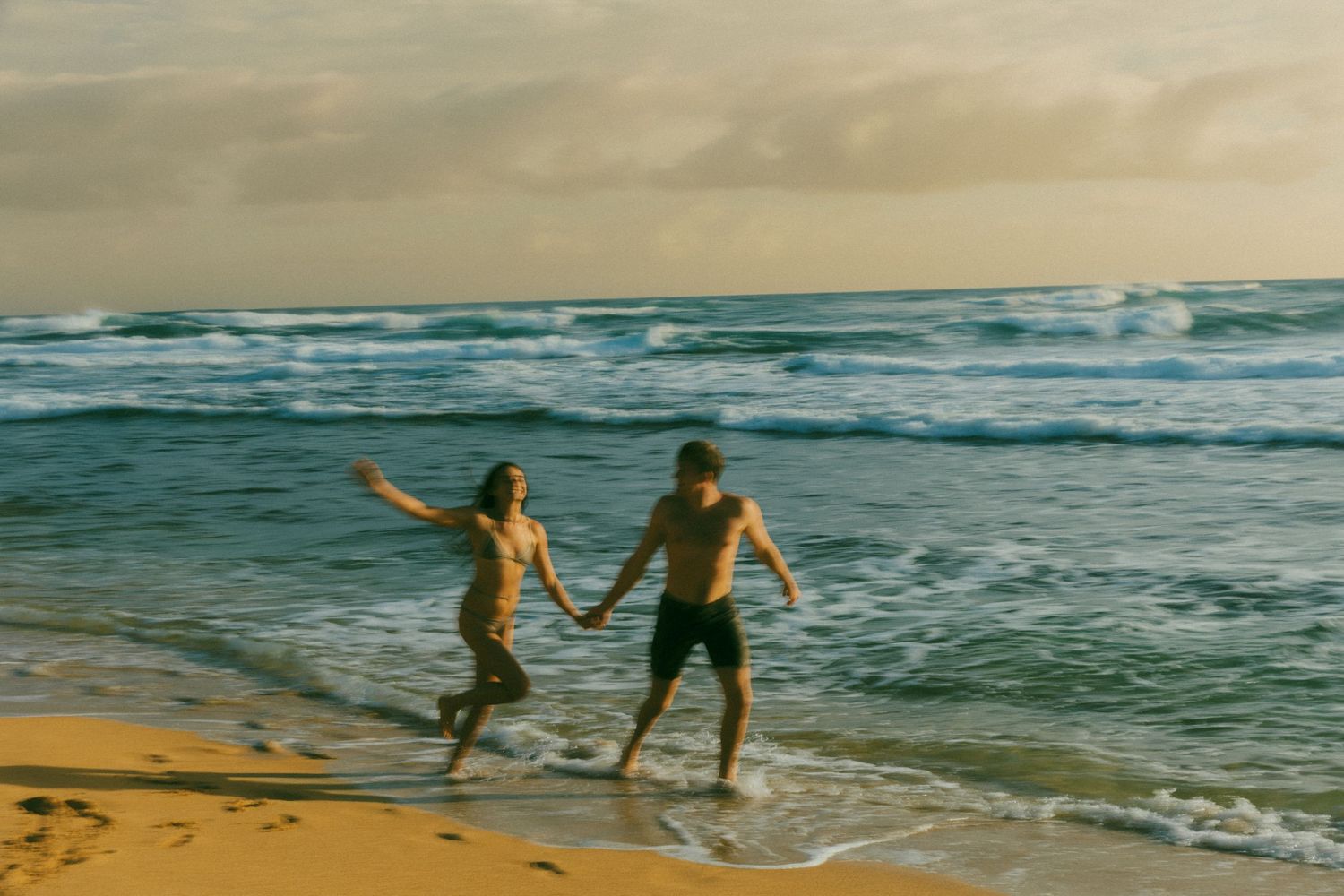 Couple holding hands running along a sandy beach with ocean waves and dramatic cloudy sky at sunset.