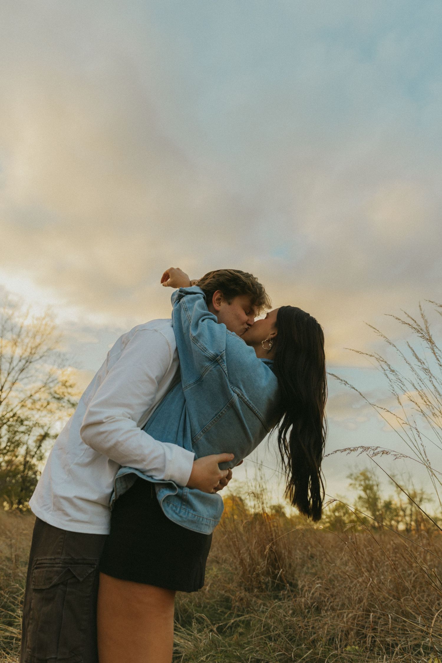 Couple sharing a romantic kiss in a golden field at sunset, embracing outdoors in casual denim and white shirt.